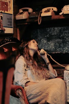 A woman leisurely sitting on a vintage chair, talking on a retro phone indoors.