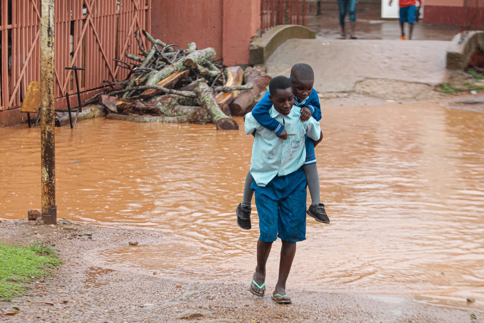 A young boy piggybacks his friend across a flooded muddy street in a village setting.