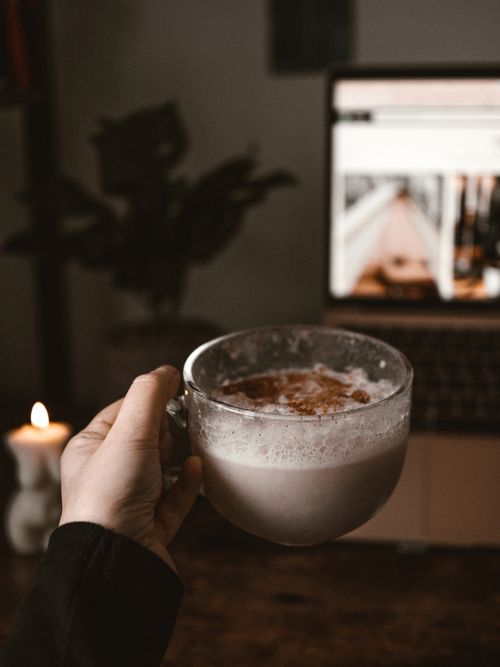 Free Hand holding a cappuccino in a cozy setting with a laptop, candle, and plants. Stock Photo
