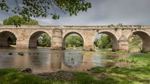 Free Ancient stone bridge arches over a calm river with lush greenery and an overcast sky. Stock Photo