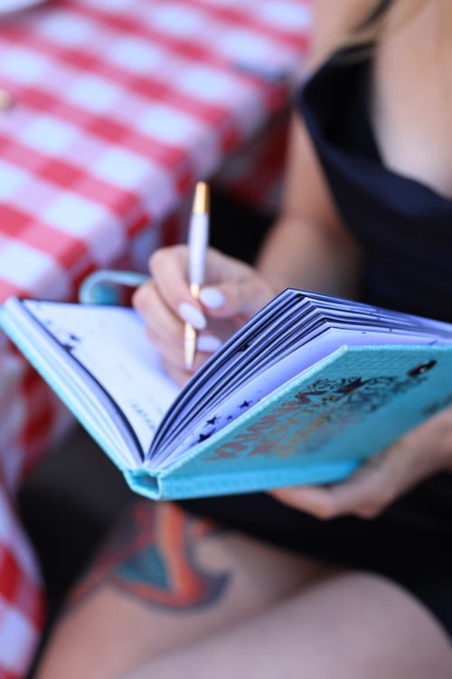 Free A woman writing in a blue notebook with a pen while sitting outdoors on a sunny day. Stock Photo