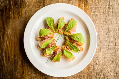 Free Delicious prosciutto on toast with leafy greens artfully arranged on a ceramic plate. Stock Photo