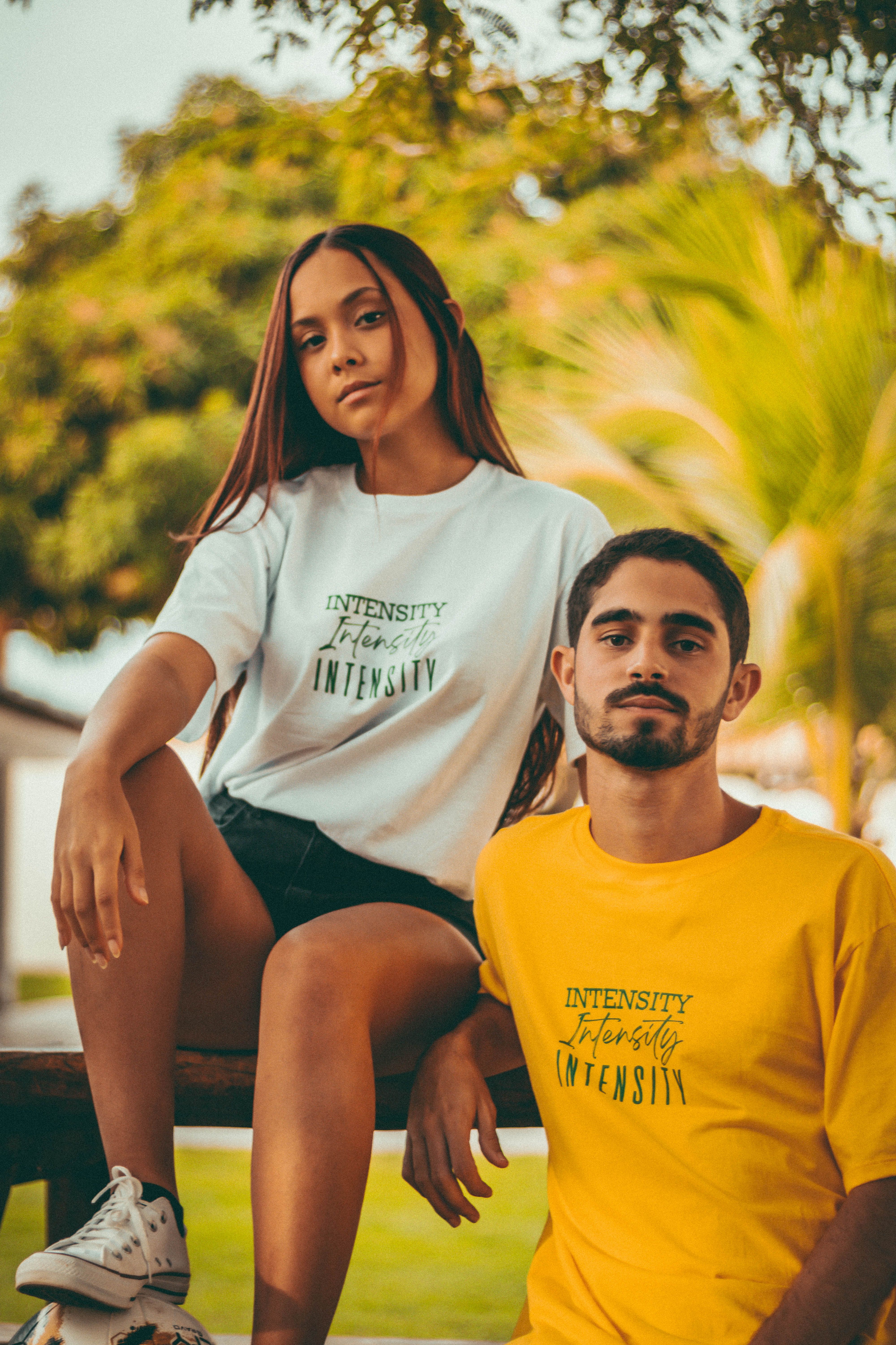 A young woman and man sitting together outdoors, showcasing trendy t-shirts with text.