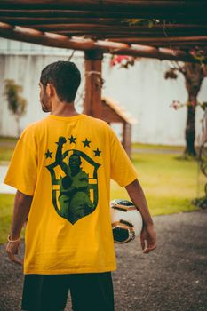 Man standing outdoors holding a soccer ball, wearing a yellow shirt with printed design.