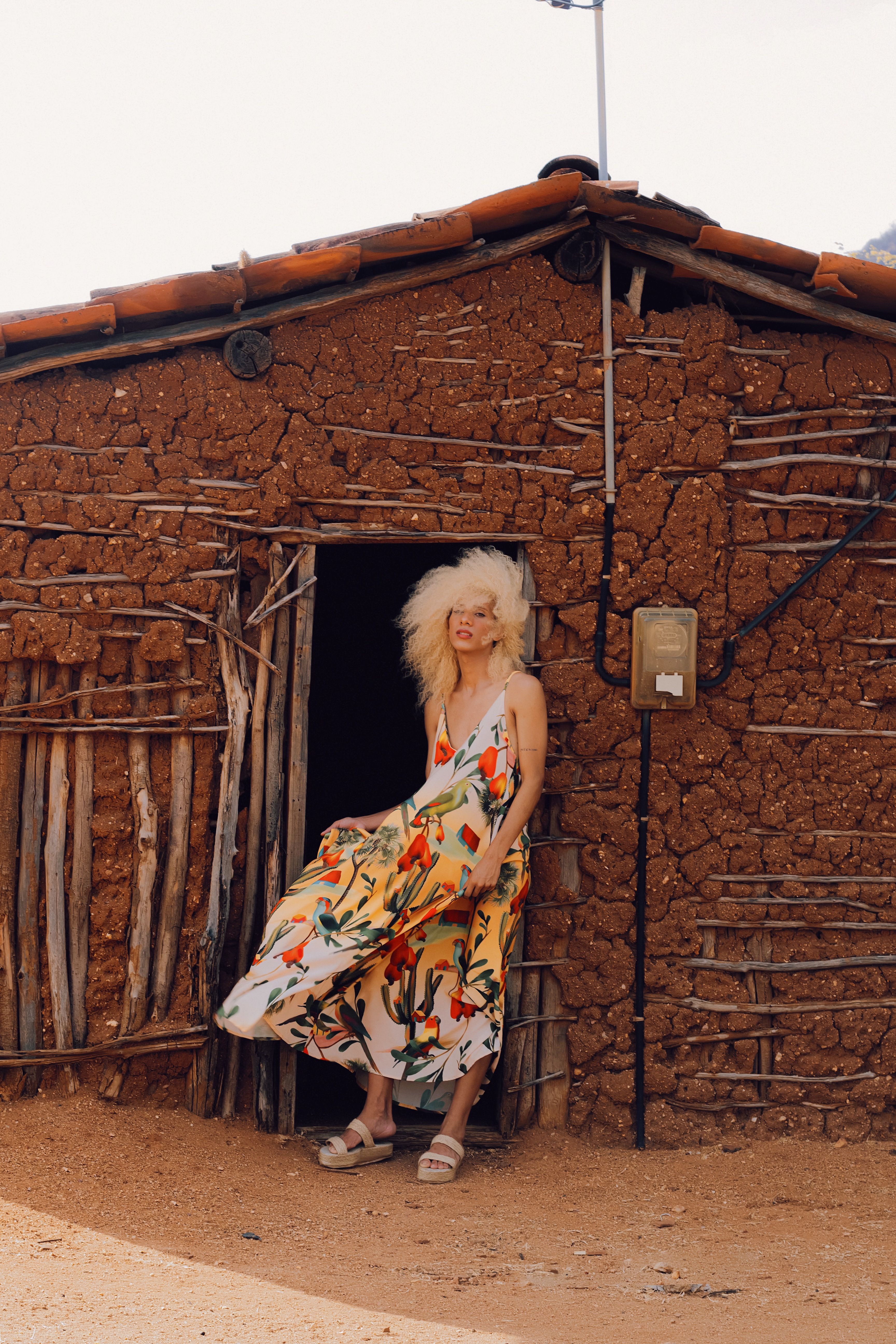 A woman in a vibrant dress poses by the entrance of a rustic hut on a sunny day.