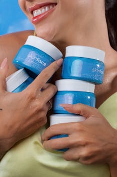 Smiling woman holding blue skincare containers against soft pastel background.