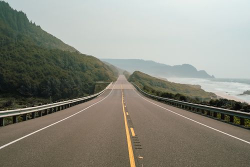 Free A tranquil view of a straight coastal road flanked by misty mountains and the ocean. Stock Photo