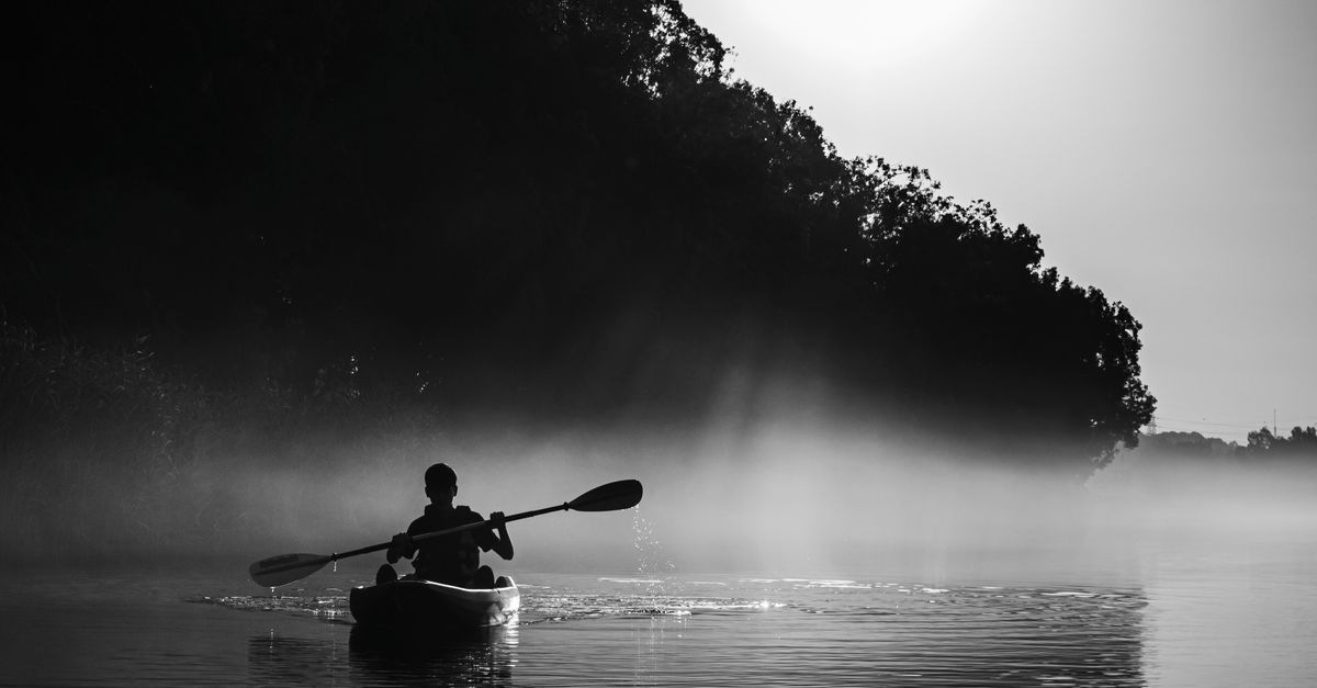 Silhouette of a man kayaking in tranquil waters with mist and morning light.