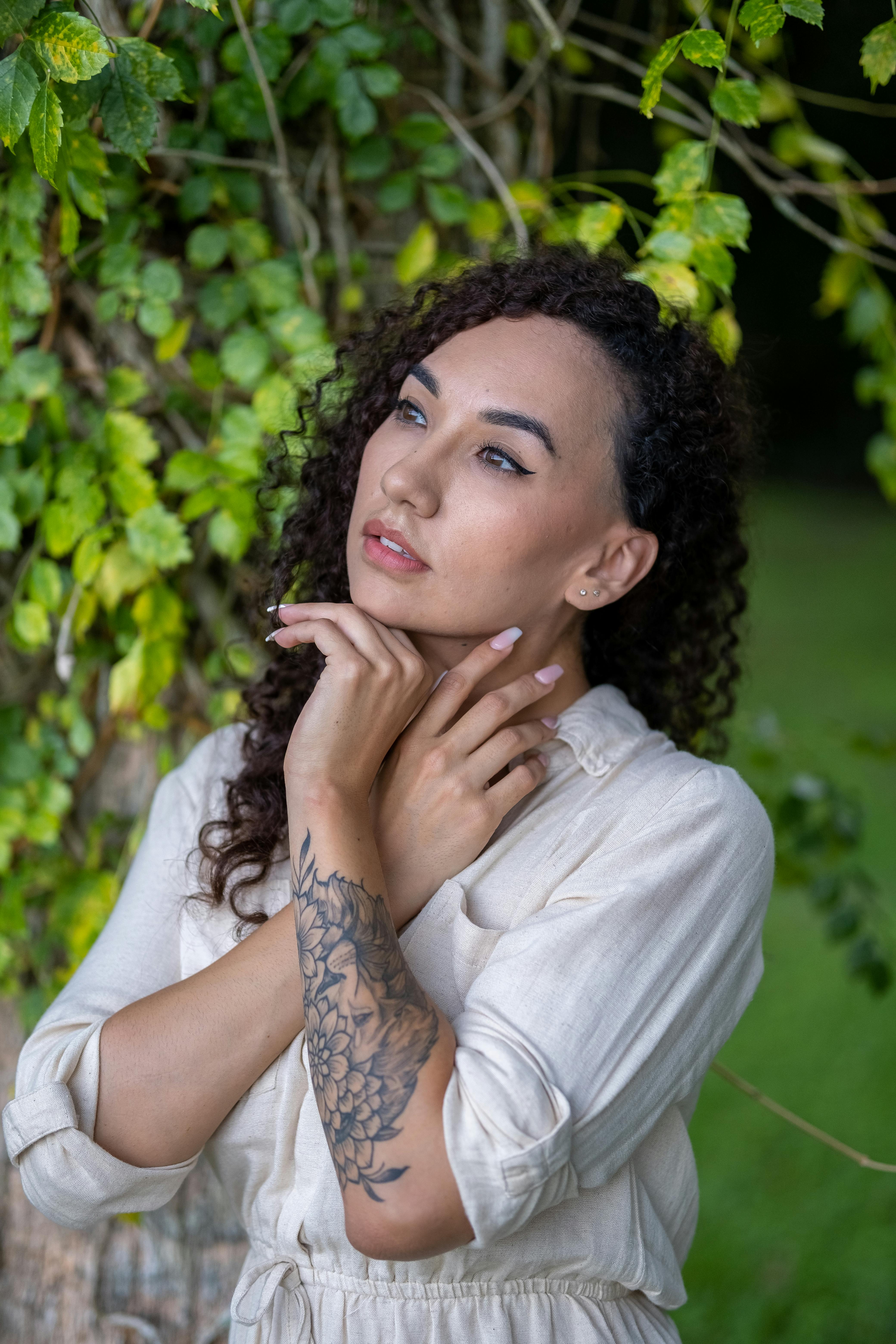 Portrait of a woman with curly hair and tattoos posing by greenery, embodying natural beauty and sty