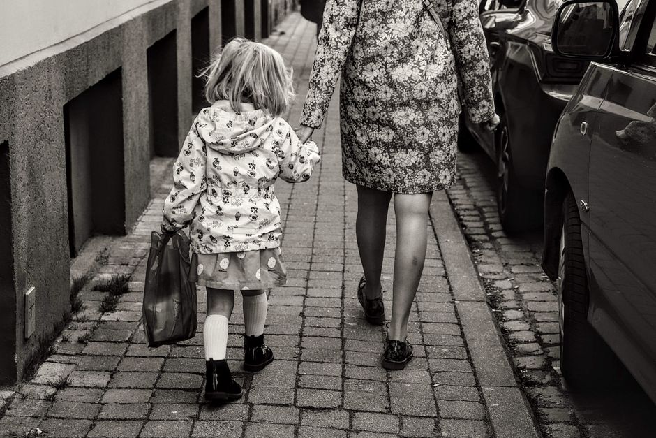 A monochrome image of a mother and daughter holding hands and walking on a city sidewalk.