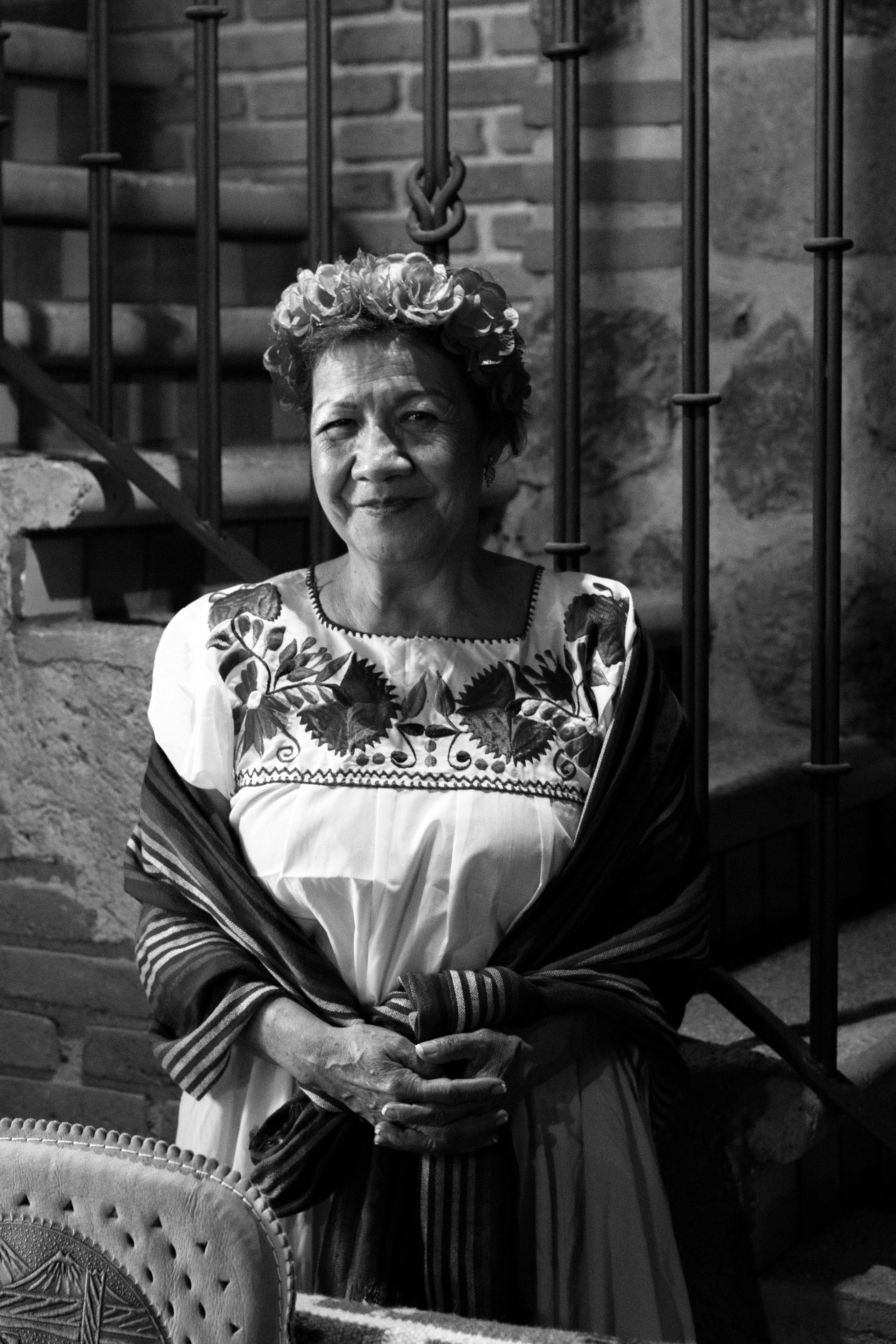 A senior woman wearing traditional Mexican dress and flower headband, captured in a grayscale portrait.