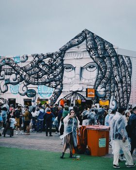 Crowd gathers at event with large mural, under cloudy skies.