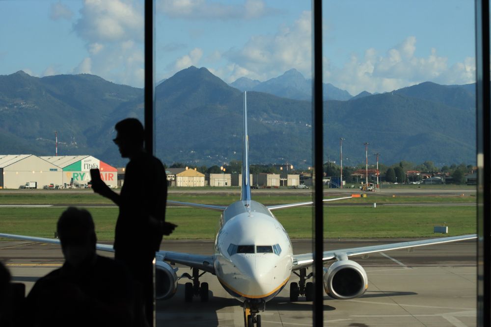 Siluetă de călător la peretele de sticlă al unui terminal de aeroport, cu avion și munți afară