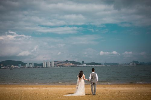Free Bride and groom holding hands on a beach, gazing at the sea under a cloudy sky. Stock Photo