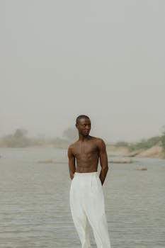 Muscular man posing topless near a serene river on a misty day, embodying strength and tranquility.