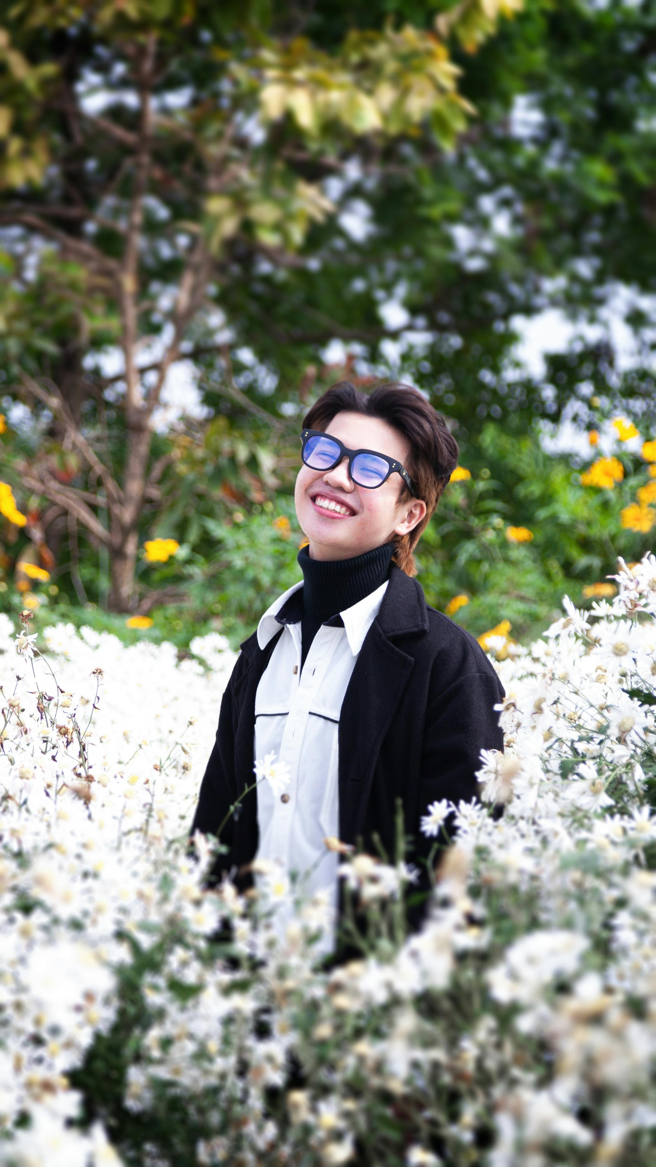 Smiling adult in a field of white flowers wearing blue glasses outdoors.