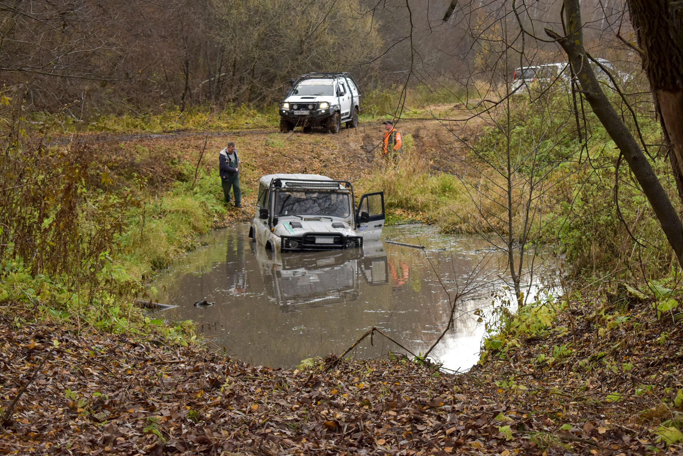 White 4x4 bogged to the hood in a muddy forest crossing with a second truck ready to perform a recovery pull