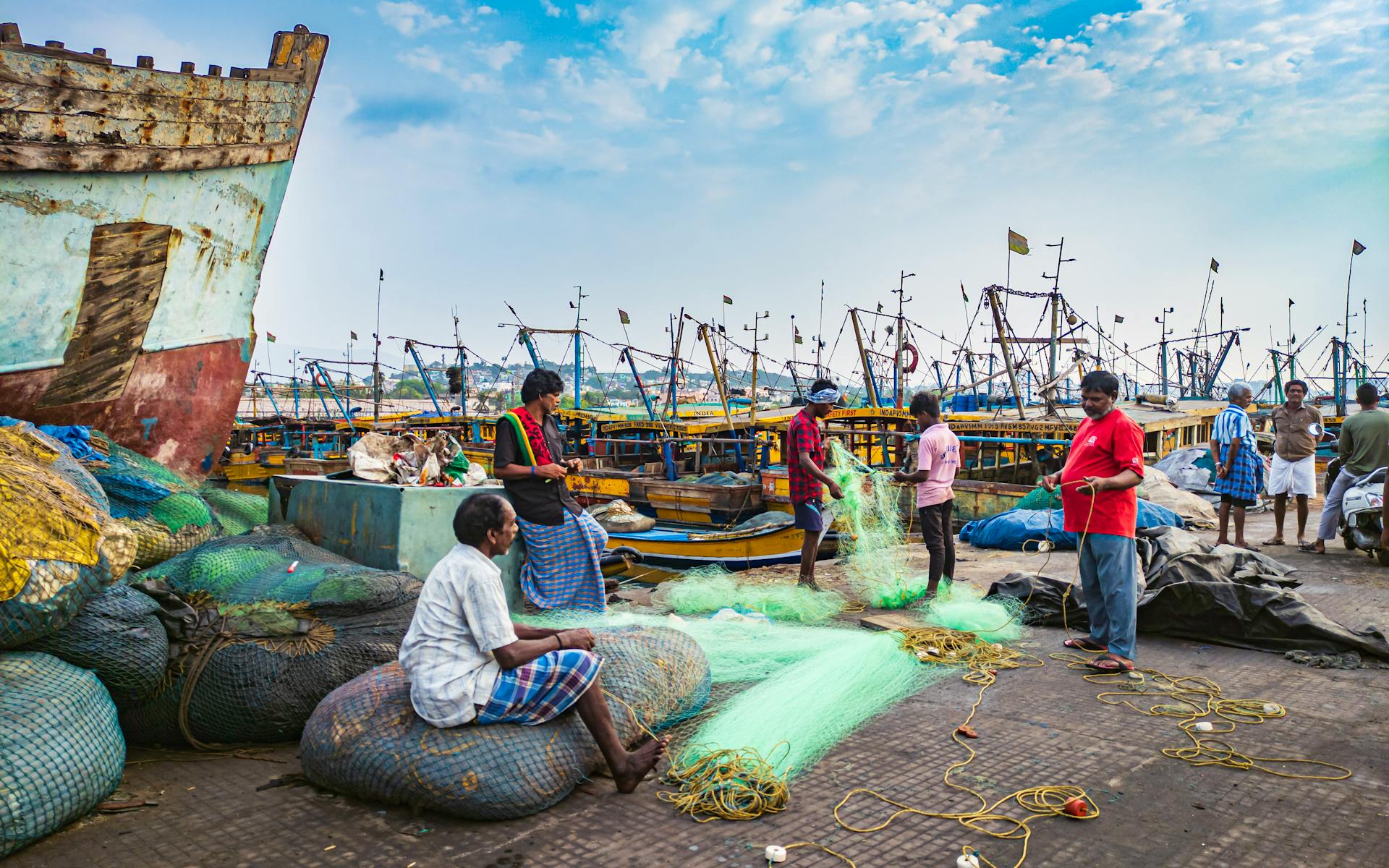 South Asian fishermen prepare nets at Visakhapatnam harbor with colorful boats in the background.