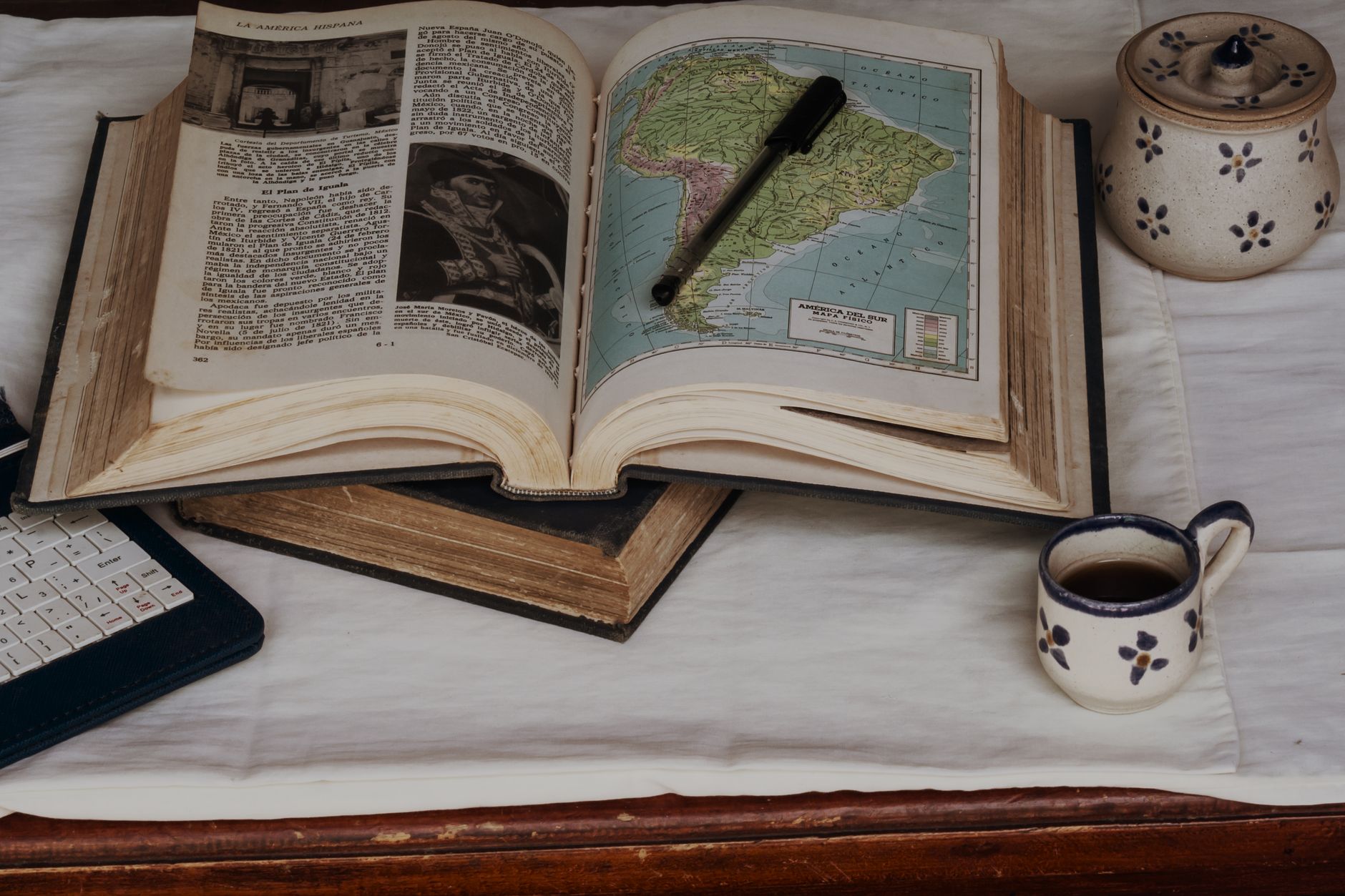 An open vintage book showing a map of South America, accompanied by a pen and a cup of coffee, evoking a scholarly atmosphere.