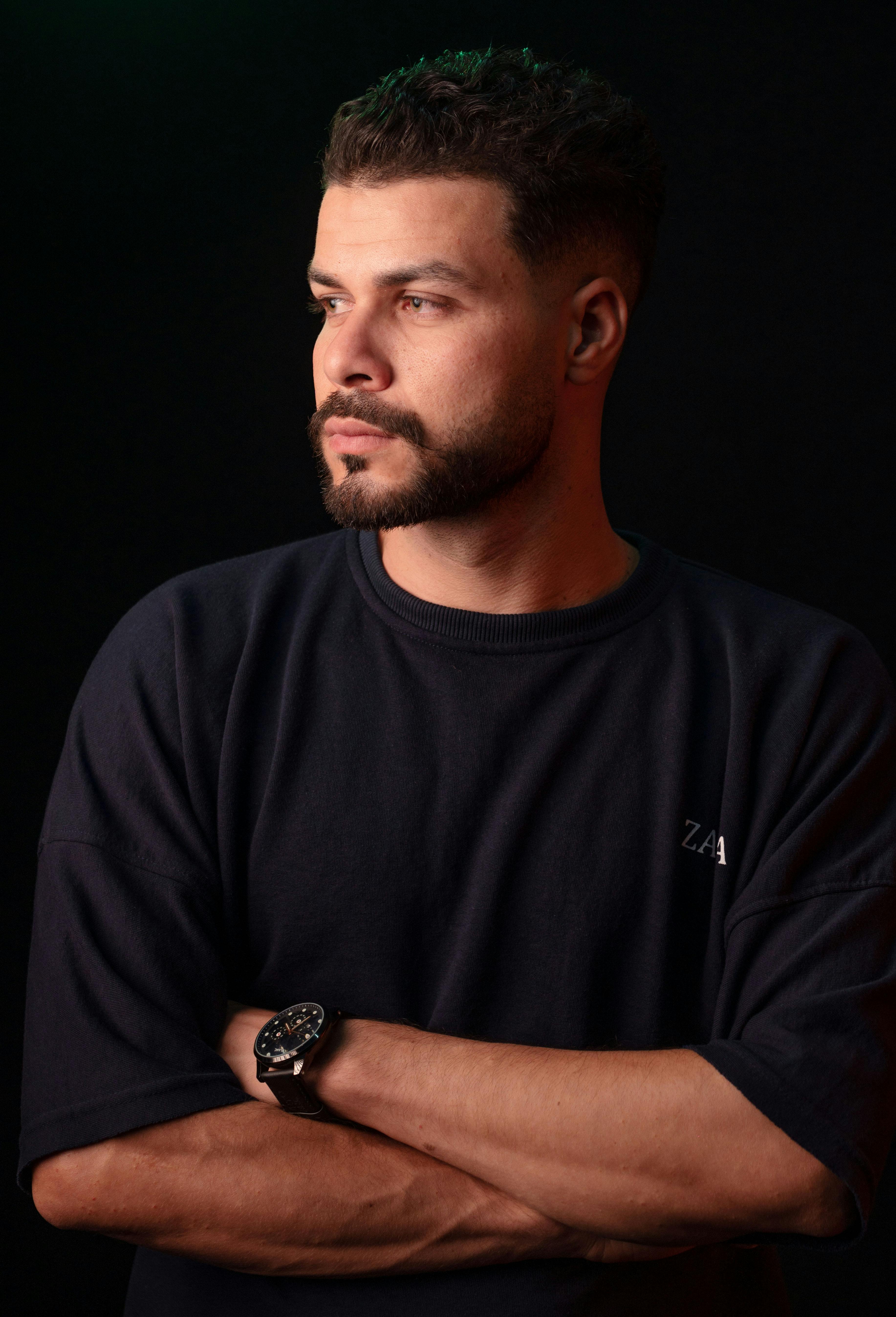Close-up of a man in a black shirt, arms crossed, against a dark background.