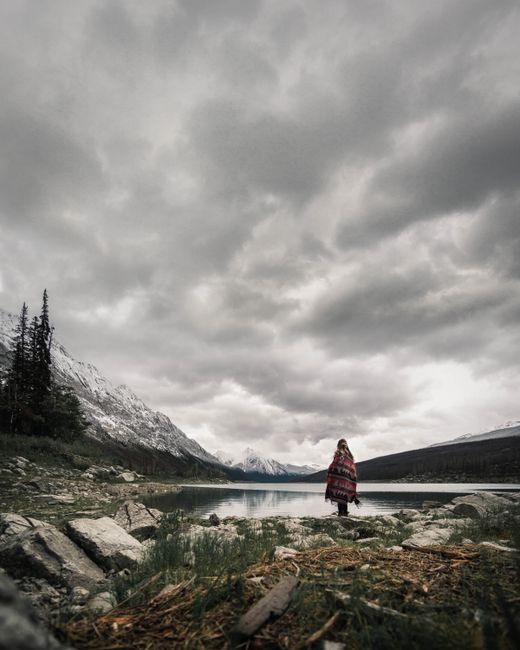 A person stands wrapped in a blanket by a serene lake, surrounded by mountains under an overcast sky.