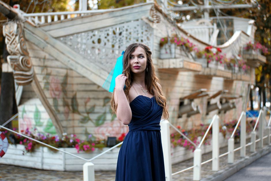Stylish woman in a blue dress stands gracefully with a whimsical wooden ship backdrop in a park.