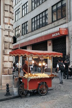 An outdoor street vendor selling corn from a red food cart in a busy urban area with people walking by.