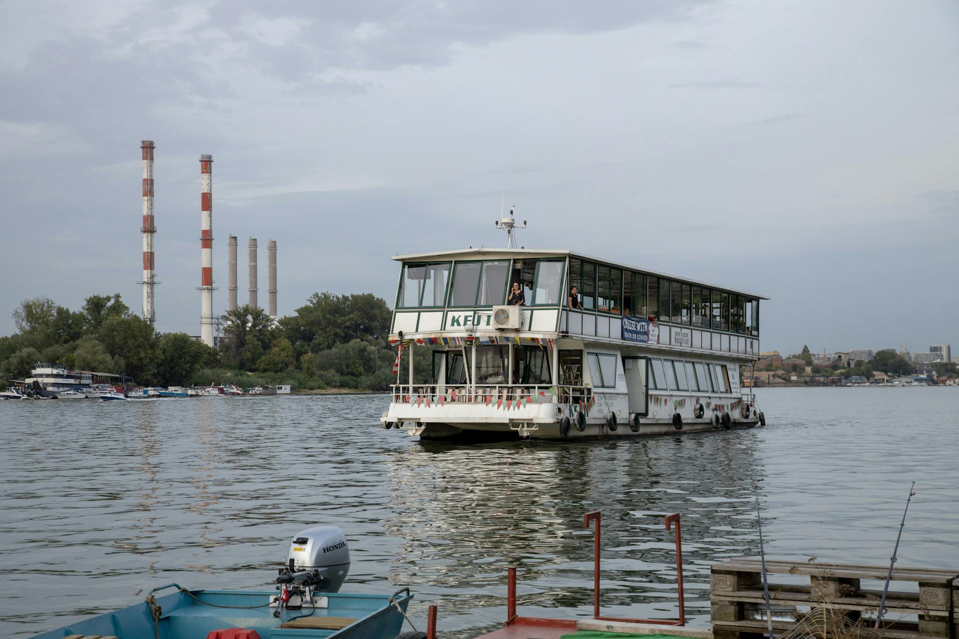 A ferry boat on a calm river with industrial chimneys in the background symbolizes transport and industry.
