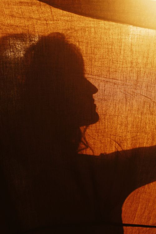 Free Artistic silhouette of a woman behind a curtain with warm sunlight creating dramatic shadows. Stock Photo