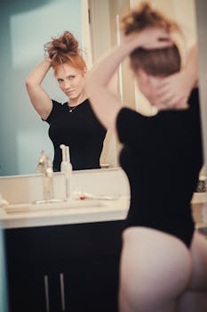 A woman in a bodysuit fixes her hair while looking in a bathroom mirror with a focused expression.
