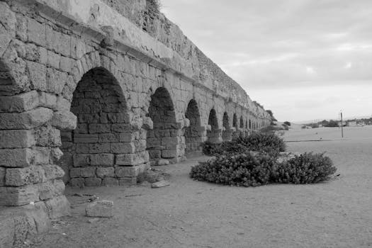 Black and white image of ancient Roman aqueduct arches on a sandy beach in Caesarea, Israel. by Lio Voo