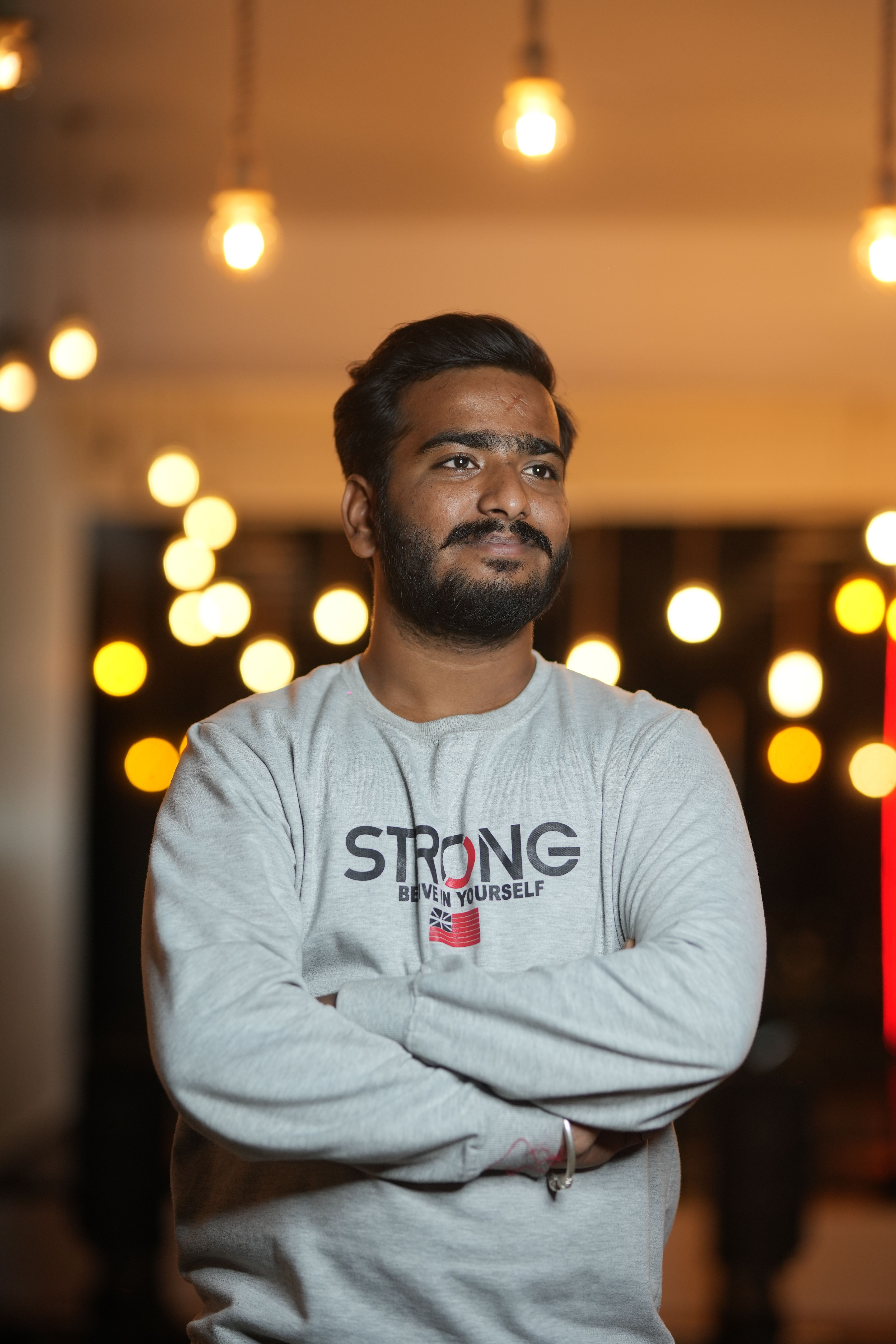 A bearded man stands confidently with arms crossed wearing a 'Strong' sweatshirt. Bright bokeh lights in the background.