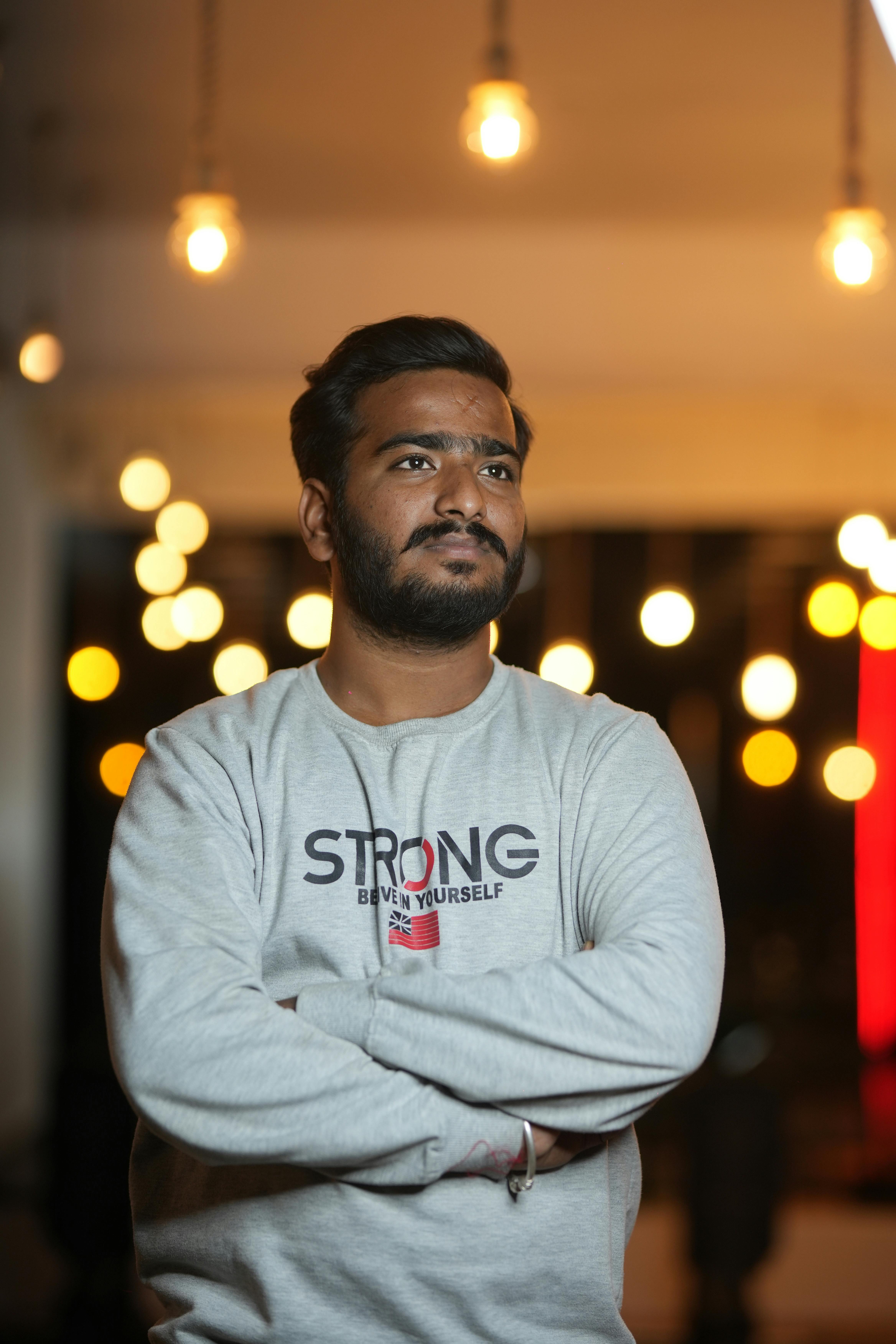 A young man stands pensive indoors, wearing a shirt with 'Strong' text, surrounded by warm lights.