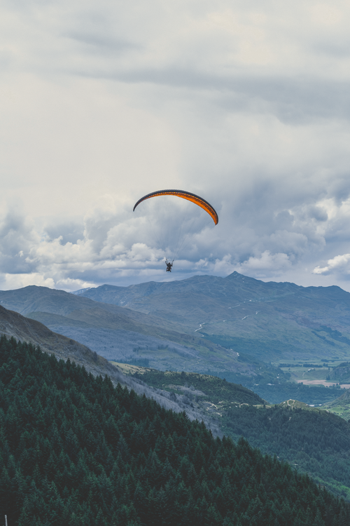 Free Scenic view of a paraglider soaring over green mountains and forests under a cloudy sky in New Zealand. Stock Photo