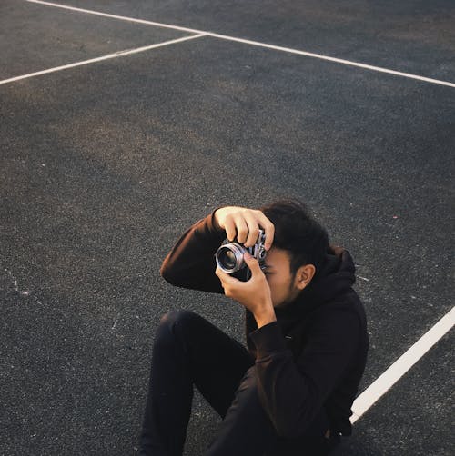 Man in Black Hoodie and Pants Sitting on Asphalt Road While Taking a Picture 