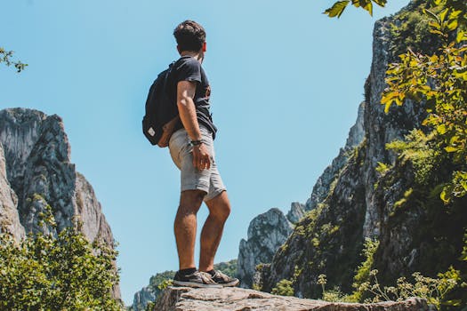 A hiker stands proudly atop a rock, overlooking a stunning mountain vista under a clear blue sky.