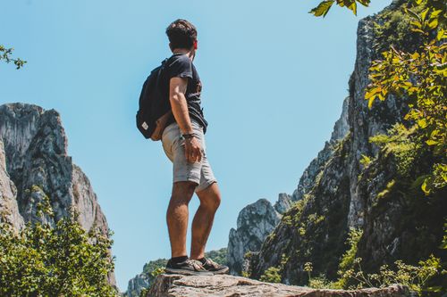 Free A hiker stands proudly atop a rock, overlooking a stunning mountain vista under a clear blue sky. Stock Photo