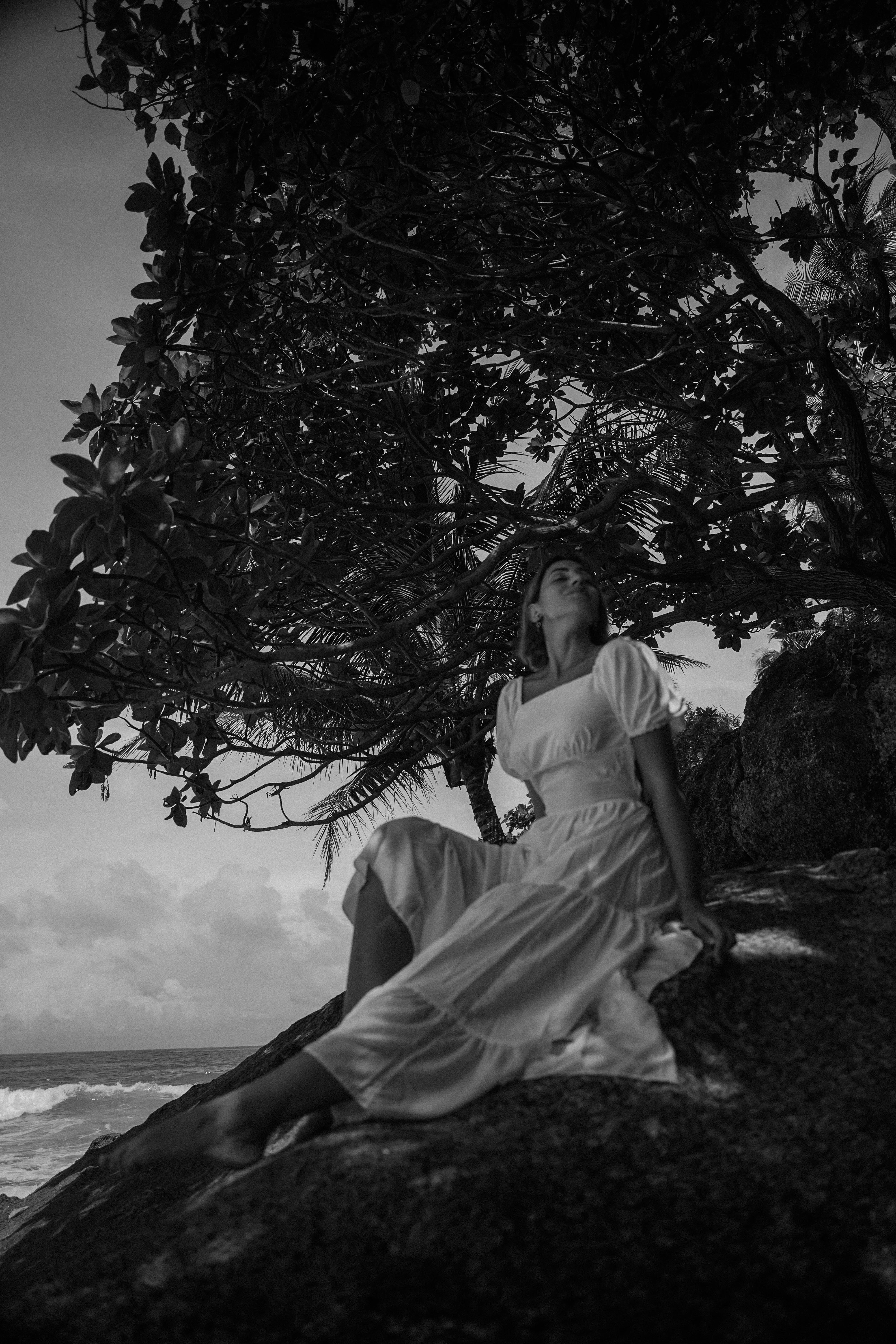 Serene scene of a woman in a white dress sitting by a tropical beach in black and white.