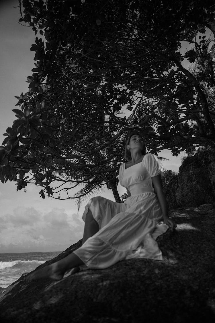 Woman Wearing A White Dress Sitting On Large Rocks On A Seashore 