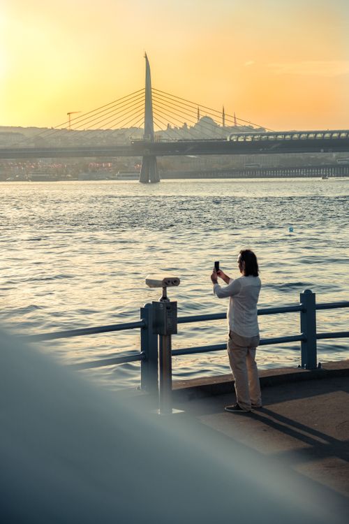 Free Man capturing sunset over Istanbul's Golden Horn Bridge on a smartphone. Stock Photo