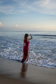 A woman stands on a sandy beach in a red dress, gazing at the serene ocean under a clear blue sky.