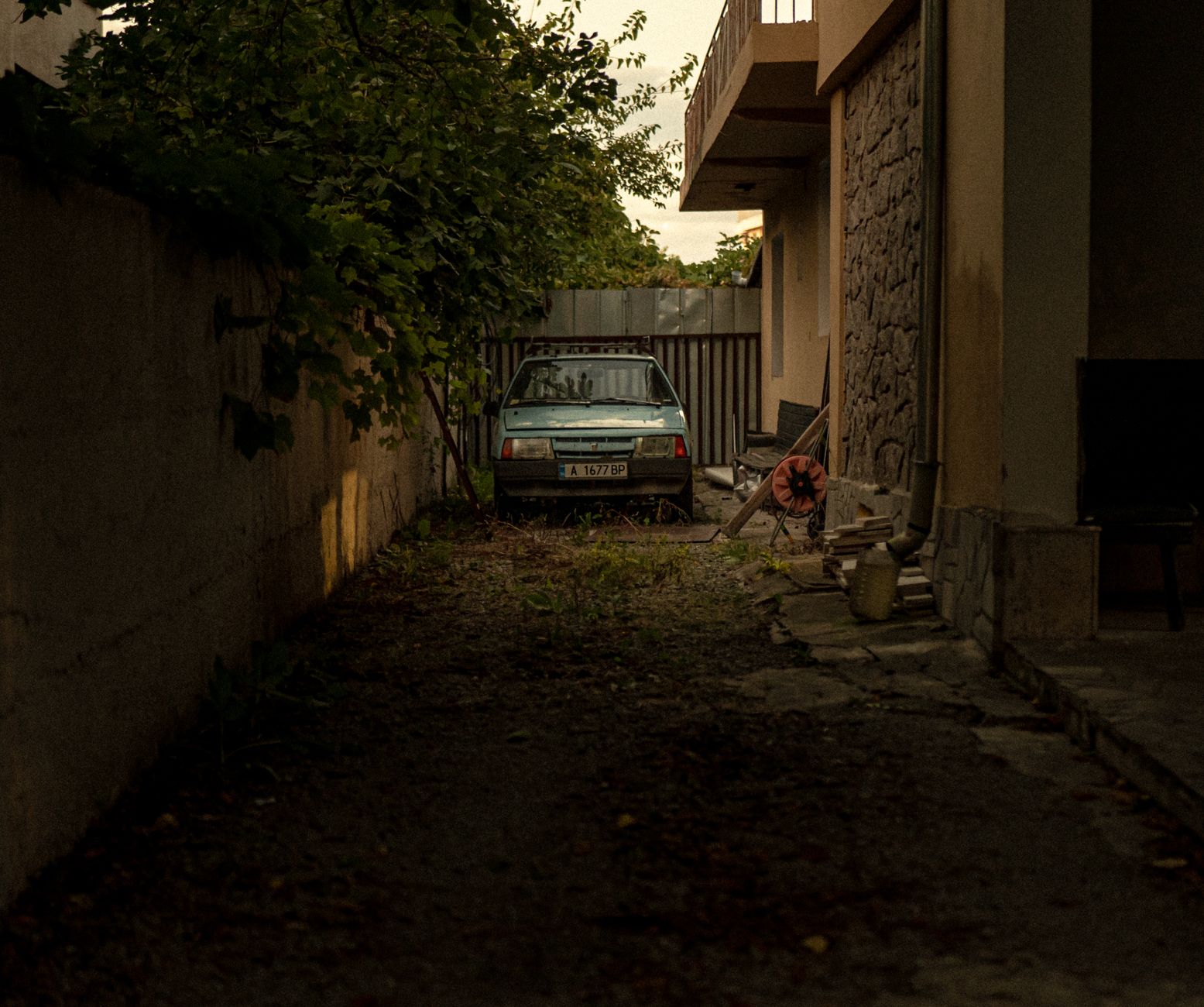 An abandoned car parked in a narrow urban alleyway, surrounded by overgrown vegetation.