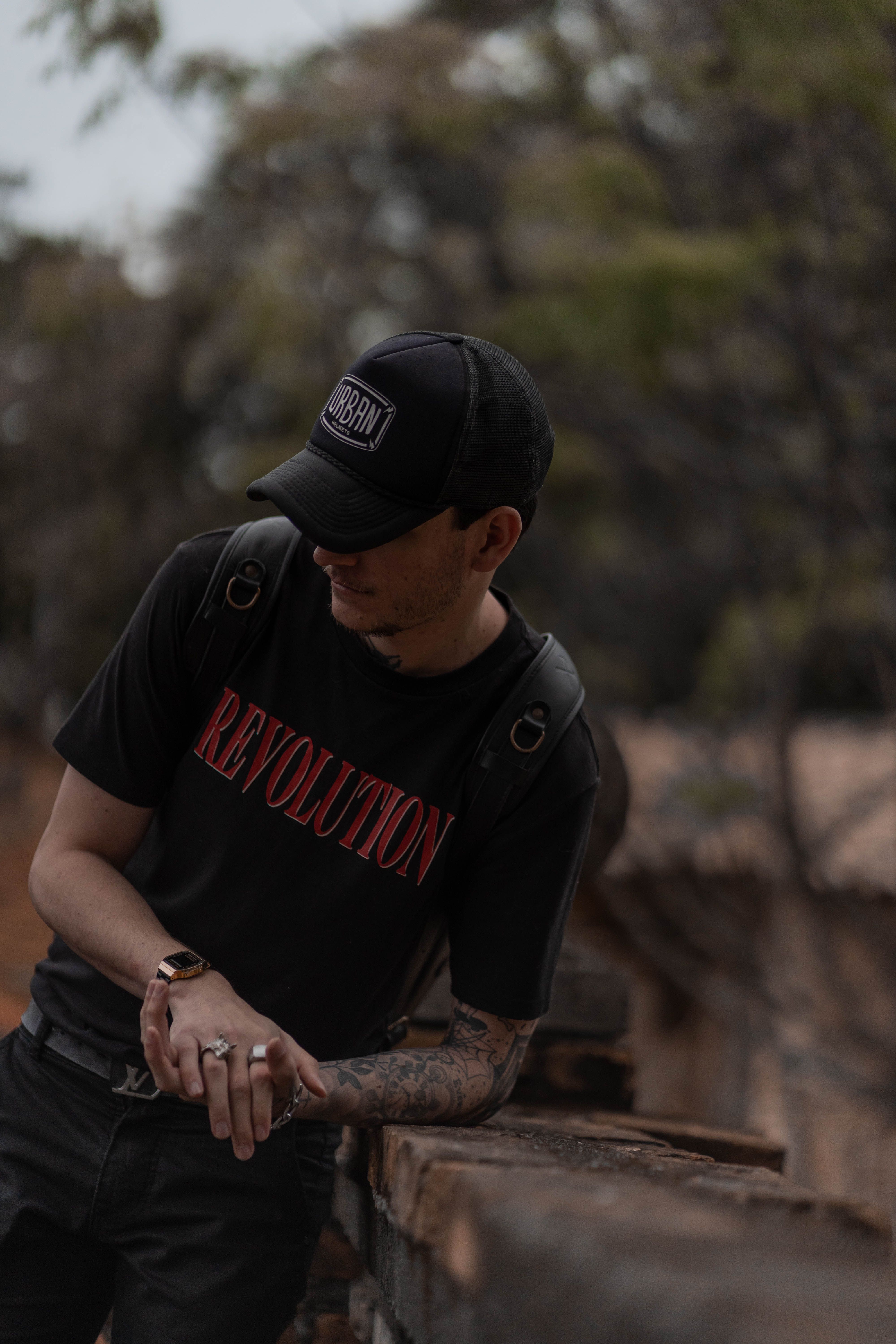Man wearing cap with arm tattoo, leaning on wall outdoors with a mysterious vibe.