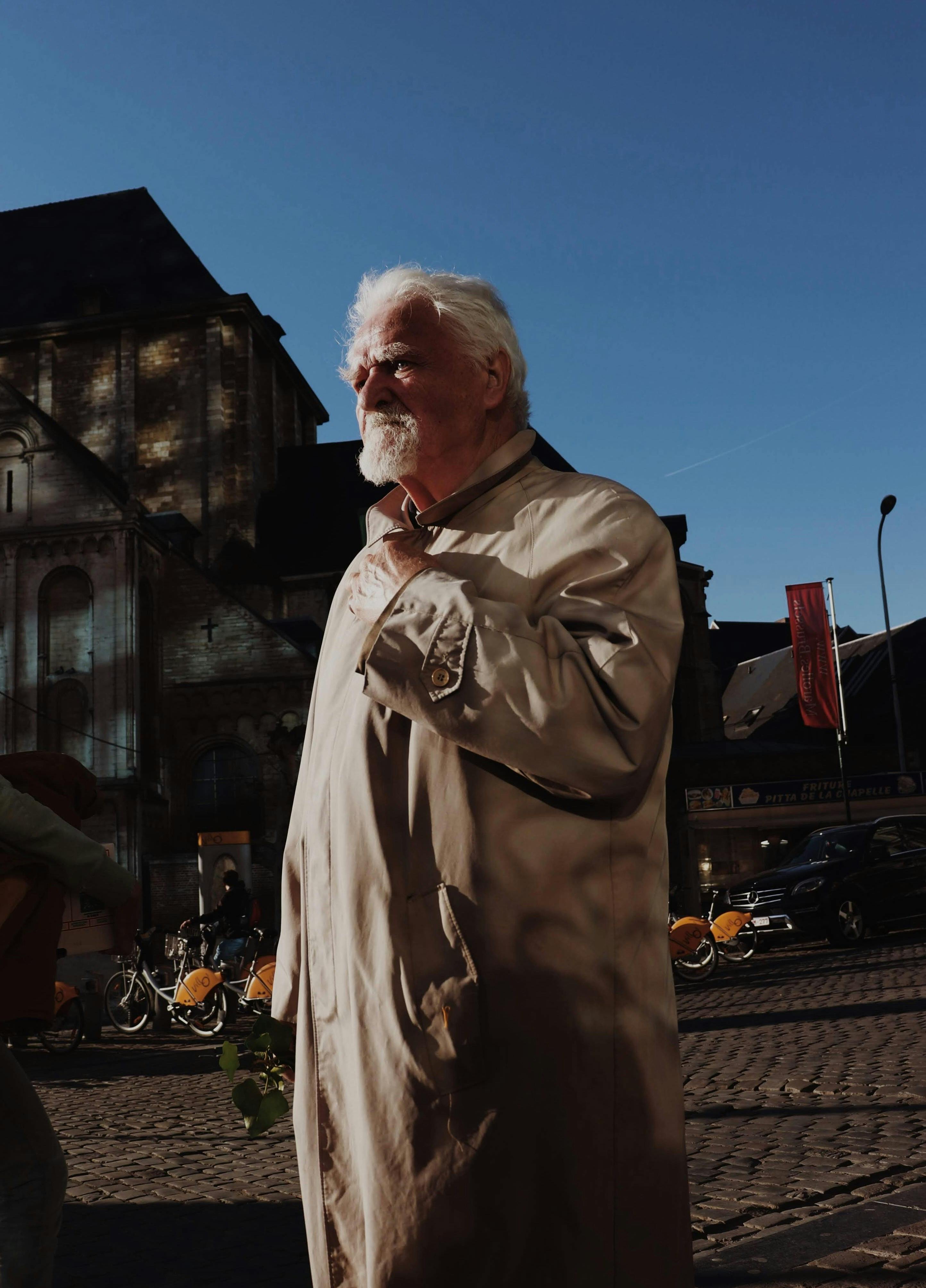 Senior man with white hair in a beige coat strolling through a sunlit street with bicycles in the background.