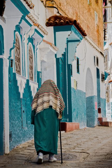 Elderly woman with shawl and cane walking along vibrant blue city street.