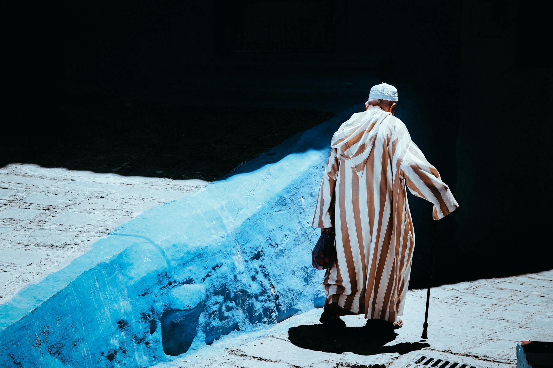 Elderly man in traditional robe walking in a sunlit Moroccan alleyway.