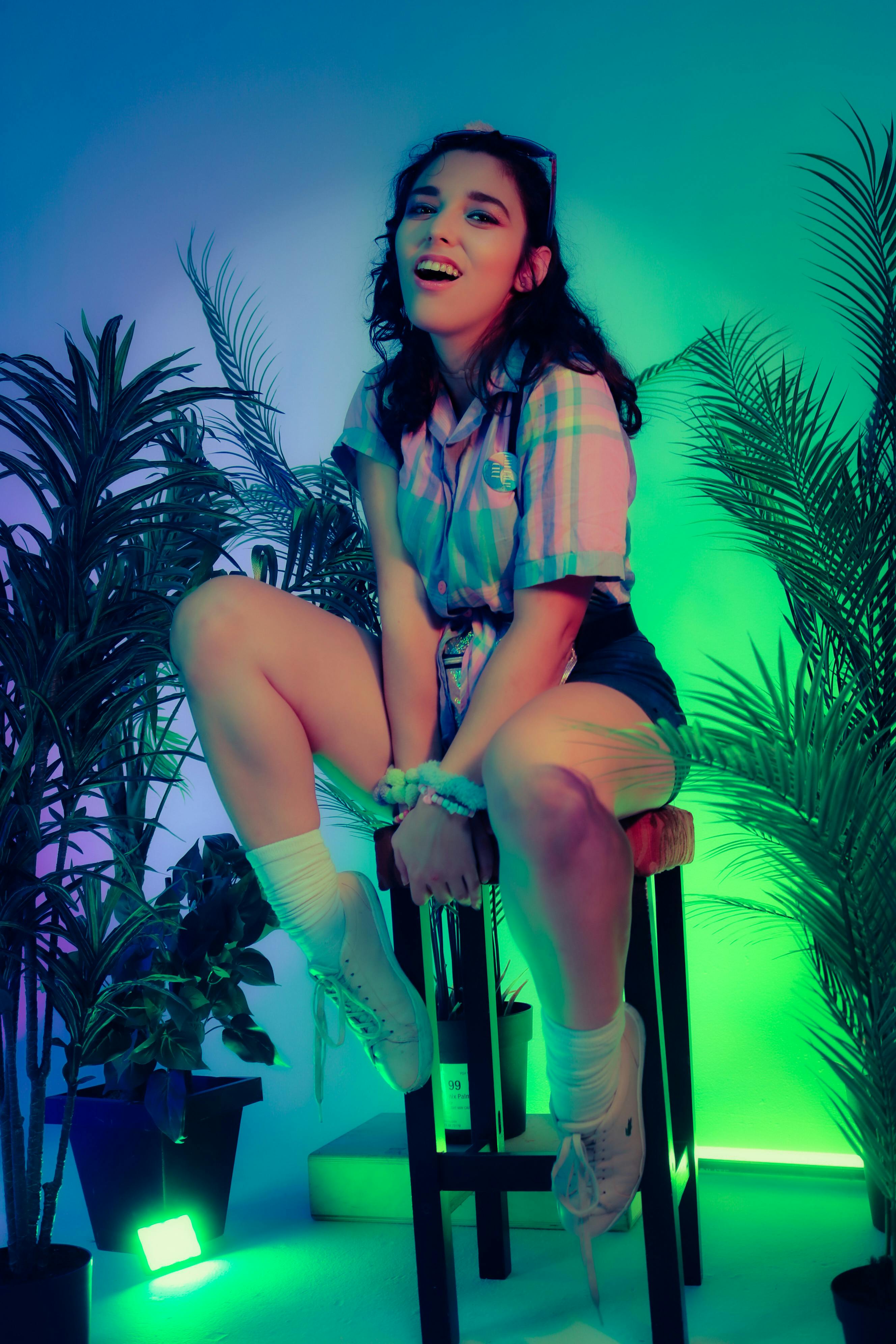 Energetic woman posing on a chair surrounded by neon lights and decorative plants in a vibrant studio setting.