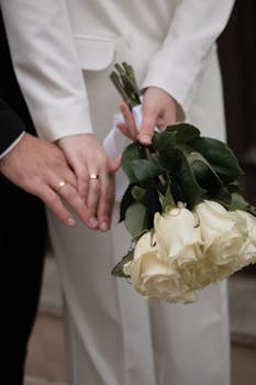 A close-up of a couple holding hands with a white rose bouquet, symbolizing love and unity at a wedding.