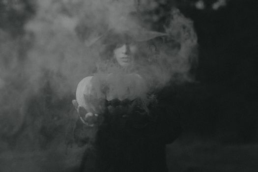 Mysterious black and white photo of a witch holding a pumpkin amidst swirling smoke, perfect for Halloween themes.