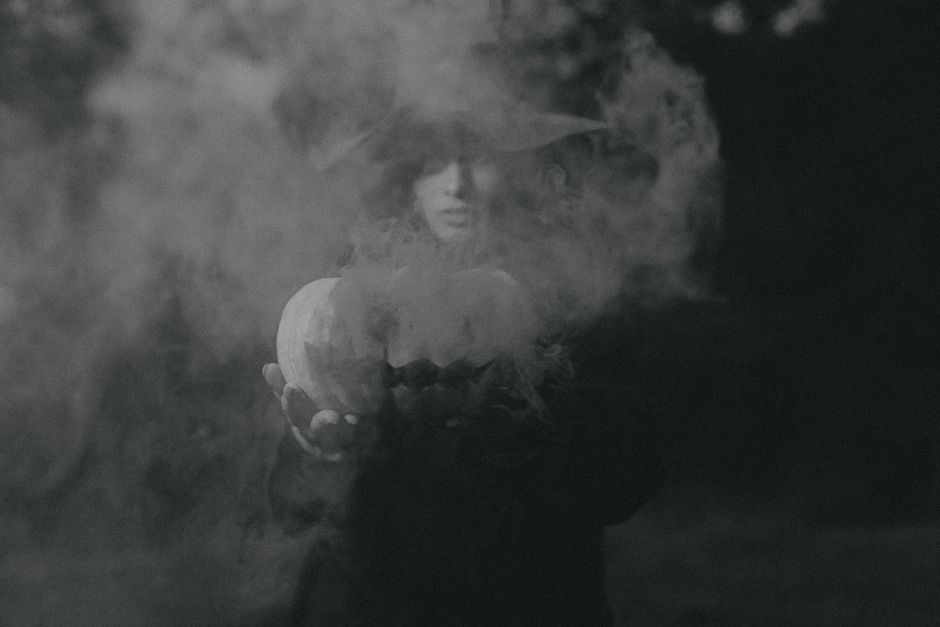 Mysterious black and white photo of a witch holding a pumpkin amidst swirling smoke, perfect for Halloween themes.
